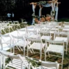 Elegancka i stylowa ceremonia ślubna w plenerze, organizowana przez RENT4WEDDING uwieczniona na zdjęciu. Białe, składane krzesła z delikatnymi dekoracjami tworzą harmonijną aleję prowadzącą do drewnianej pergoli weselnej, udekorowanej białymi, zwiewnymi tkaninami i świeżymi kwiatami. W tle majestatyczny las stanowi idealne tło dla tego wyjątkowego dnia. Wydarzenie to doskonale oddaje kunszt i dbałość o detale rent4weddings.pl, gdzie możesz wypożyczyć wszystko, czego potrzebujesz na swoje wesele w Wrocławiu i okolicach - rent4events.pl wypożycz wrażenia, rent4wedding.pl zachowaj wspomnienia.