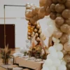Dekoracja stołu w stylu boho z plecionymi poduszkami jako siedziska oraz eleganckim stołem pokrytym beżowym, camelowy, welurowym obrusem. Stół udekorowany jest suszonymi roślinami, złotymi liśćmi oraz zielonymi szklankami. Wokół stołu rozciągają się dekoracyjne aranżacje balonowe w odcieniach złota, brązu, beżu i perłowej bieli. Scenografia jest idealna na ślub, wesele lub event, oferowana przez wypożyczalnię rent4events.pl we Wrocławiu, która specjalizuje się w scenografiach o naturalnym i eleganckim charakterze - rent4events.pl wypożycz wrażenia, rent4weddings.pl zachowaj wspomnienia.