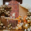 Dekoracja słodkiego stołu w stylu Boho Safari, z dominującymi balonami w odcieniach złota, brązu i perłowej bieli. W tle znajduje się ścianka w motyw żyrafy, który wzmacnia stylistykę Safari. Dwie złoto-szklane konsole udekorowane są złotymi i drewnianymi akcesoriami, na którym umieszczone są słodkie przekąski i ozdoby. Balony w kształcie łuku otaczają przestrzeń, a w rogu widać figurkę geparda. Całość tworzy elegancką i egzotyczną scenografię, idealną na ślub, wesele lub event we Wrocławiu. Wypożyczalnia dekoracji rent4events.pl zapewnia pełne wyposażenie w stylu Safari - rent4events.pl wypożycz wrażenia, rent4weddings.pl zachowaj wspomnienia.