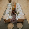 Stół w stylu Safari Boho, udekorowany bieżnikiem z motywem skóry żyrafy, suszonymi roślinami oraz elegancką zastawą stołową. Plecione Siedziska Boho z juty i bawełnianego obszycia w stylu boho leżą dookoła stołu, a zielone kieliszki i talerze z roślinnymi wzorami podkreślają naturalny, egzotyczny charakter dekoracji. Aranżacja doskonała na eventy, śluby oraz wesela organizowane we Wrocławiu i okolicach. Wypożyczalnia rent4events.pl oferuje szeroki wybór dekoracji, łączących styl safari z boho, tworząc oryginalne i eleganckie scenografie - rent4events.pl wypożycz wrażenia, rent4weddings.pl zachowaj wspomnienia