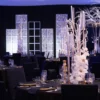 Wypożyczalnia dekoracji Rent4Events prezentuje elegancką zimową aranżację eventową sali. Na zdjęciu widoczne są ozdobione stoły z wysokimi świecznikami, białymi poinsecjami i gałęziami, podkreślonymi złotymi akcentami. Tło tworzą podświetlane panele świetlne, które dodają przestrzeni romantycznego nastroju. W głębi sali znajduje się srebrna figura jelenia, nadająca całości zimowego charakteru. Scenografia idealnie sprawdzi się na imprezy świąteczne i Winter Party we Wrocławiu - rent4events.pl wypożycz wrażenia, rent4wedding.pl zachowaj wspomnienia. Fot. @karolinakiec_fotografia.
