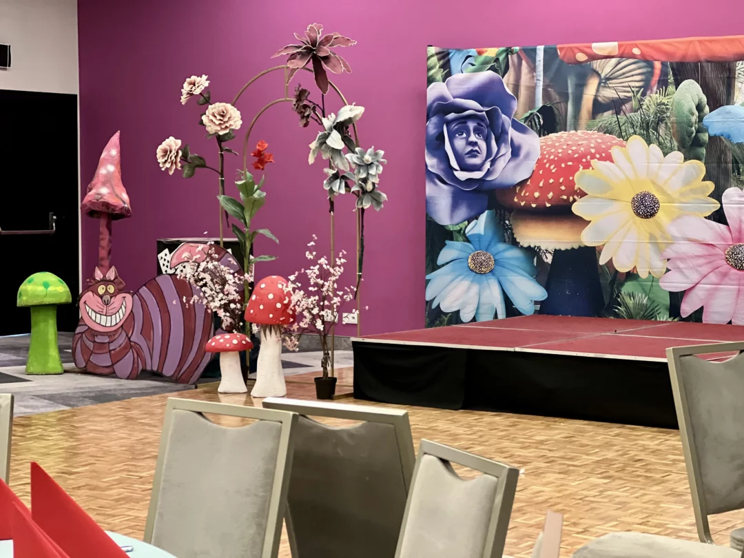 Fantazyjna dekoracja inspirowana „Alicją w Krainie Czarów”, z szeroko uśmiechniętym fioletowym Kotem z Cheshire na tle różowych i zielonych grzybów. Scenografia ozdobiona jest ogromnymi, kolorowymi kwiatami upiętymi na złotym łuku, oraz tłem fotograficznym (Horyzont Alicja w Krainie Czarów Grzyby 3) z niebieskim kwiatem z twarzą, innymi kwiatami i grzybami. Scena i podest tworzą przestrzeń, która jest idealna na baśniowy event we Wrocławiu. Dekoracje dostępne są w wypożyczalni rent4events.pl - rent4events.pl wypożycz wrażenia, rent4wedding.pl zachowaj wspomnienia. Fot. Jona.