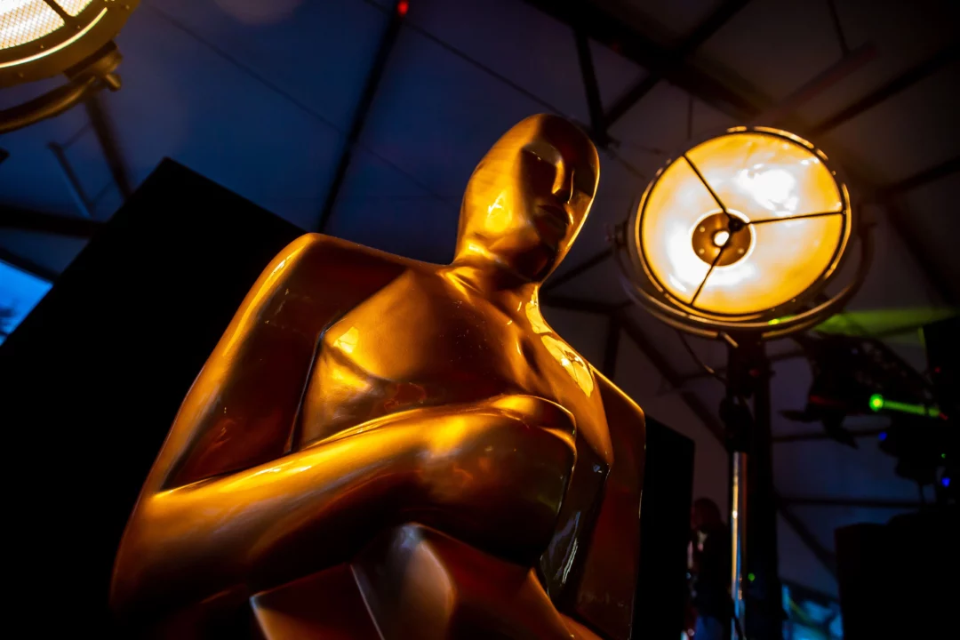 Zbliżenie na złotą statuetkę Oscara o gładkiej i błyszczącej powierzchni, oświetloną przez profesjonalny reflektor emitujący żółto-pomarańczowe światło. Nowoczesna aranżacja dekoracji na ciemnym tle, podkreślająca prestiżowy charakter scenografii. Połączenie światła i luksusowej stylistyki doskonale oddaje atmosferę ekskluzywnych wydarzeń organizowanych z pomocą wypożyczalni Rent4Events.pl i Rent4Weddings.pl. Ta aranżacja dekoracji scenicznych wyróżnia się estetyką i klasą - rent4events.pl wypożycz wrażenia, rent4weddings.pl zachowaj wspomnienia. Fot. Powerevents @powerevents.
