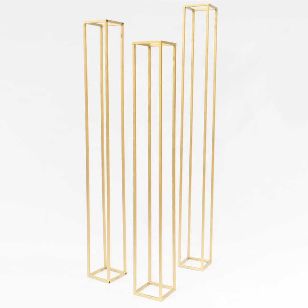 Trzy Złote Słupy o różnych wysokościach. Zestaw trzech złotych słupków dekoracyjnych o wysokościach 225 cm, 205 cm i 250 cm, z podstawą 25x25 cm. Wykonane z metalu o minimalistycznym, geometrycznym designie, idealnie komponują się w stylu glamour i romantycznym. Doskonałe do kompozycji kwiatowych i dekoracji przestrzeni podczas ślubów, wesel i eventów. Złoty kolor dodaje elegancji i prestiżu aranżacjom. Wynajem dostępny we Wrocławiu – rent4events.pl i rent4weddings.pl - rent4events.pl wypożycz wrażenia, rent4weddings.pl zachowaj wspomnienia.