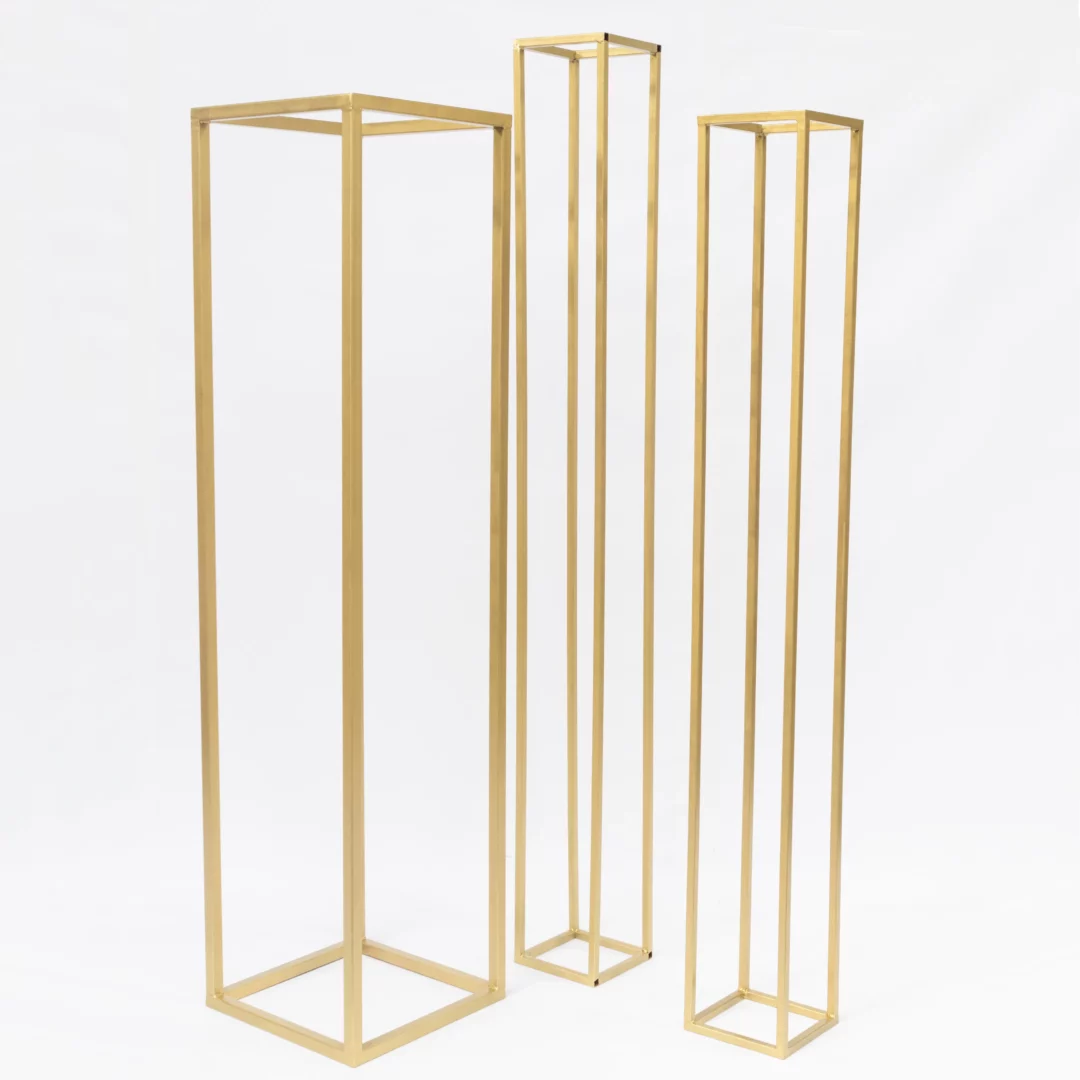 Trzy Złote Słupy o różnych wysokościach. Zestaw trzech złotych słupków dekoracyjnych o wysokościach 225 cm, 205 cm i podstawach 25 x 25 cm oraz o wysokości 206 cm i podstawie 50 x 50 cm. Minimalistyczna, geometryczna forma wykonana z profilu stalowego doskonale pasuje do stylu glamour i romantycznego. Idealne do kompozycji kwiatowych i dekoracji podczas ślubów, wesel oraz eventów. Złoty kolor dodaje elegancji i prestiżu każdej aranżacji. Dostępne w wypożyczalni dekoracji we Wrocławiu – rent4events.pl i rent4weddings.pl - rent4events.pl wypożycz wrażenia, rent4weddings.pl zachowaj wspomnienia.