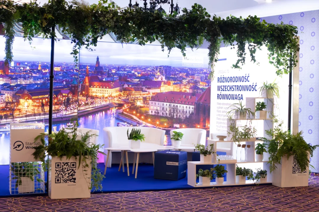 Stoisko eventowe „Wrocław – miasto spotkań” przygotowane przez wypożyczalnię rent4events.pl / rent4weddings.pl. Eleganckie białe meble, zielone rośliny w donicach, wiszące dekoracje i tło z panoramą miasta tworzą wyjątkową scenografię. Wypożyczalnia dekoracji oferuje kompleksowe wyposażenie i aranżacje, które podnoszą prestiż każdej uroczystości. Dla organizatorów ślubów, wesel i eventów we Wrocławiu i okolicach rent4events.pl i rent4weddings.pl to gwarancja stylu i jakości - rent4events.pl wypożycz wrażenia, rent4weddings.pl zachowaj wspomnienia. Fot. @karolinakiec_fotografia.