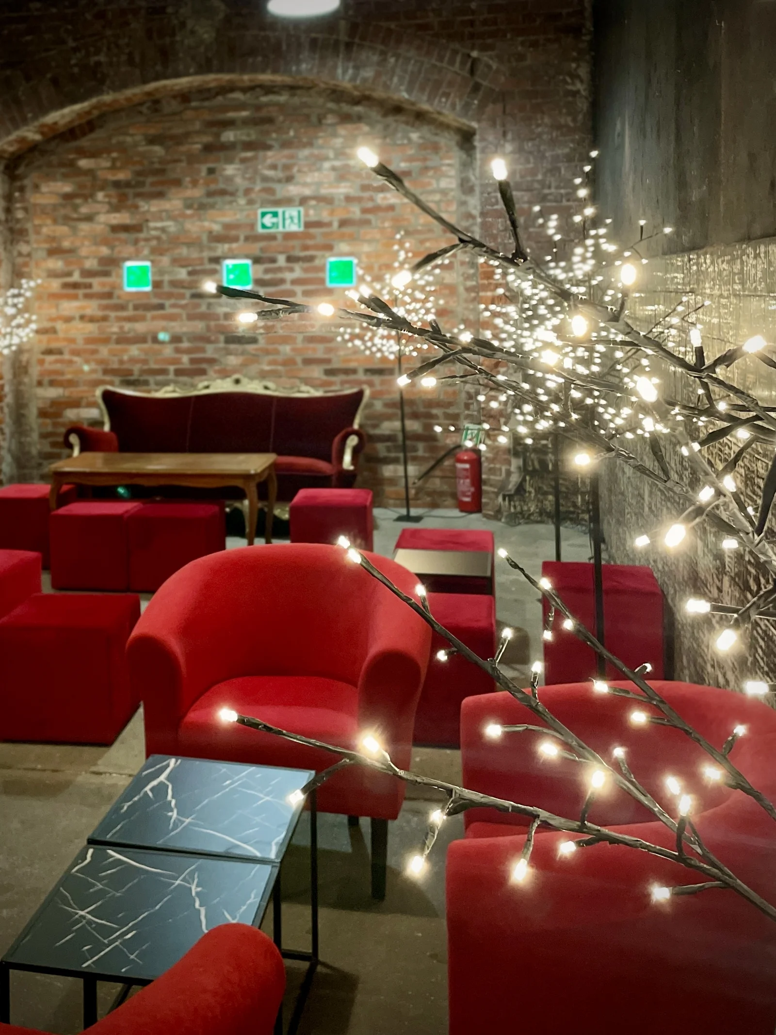aranzacja strefa wypoczynku mikolajki swieta czarne stoliki czerwone fotele drzewka led eventy wypozyczalnia dekoracji rent4events rent4wedding wroclaw - Wypożyczalnia Ślubna Czerwony chillout i Drzewka Led. Industrialna przestrzeń eventowa z ceglaną ścianą w tle. Elegancka czerwona sofa i fotele o aksamitnej tapicerce kontrastują z betonowym podłożem. W otoczeniu znajdują się sześcienne, czerwone pufy oraz nowoczesne, czarne stoliki. Dekoracyjne drzewka LED z delikatnym oświetleniem tworzą magiczną atmosferę. Industrialne akcenty, jak oświetlenie sufitowe i surowa, ceglana struktura, nadają wnętrzu unikalny charakter. Wypożyczalnia dekoracji i scenografii na event, ślub oraz wesele we Wrocławiu - rent4events.pl wypożycz wrażenia, rent4weddings.pl zachowaj wspomnienia. Fot. Matyna.