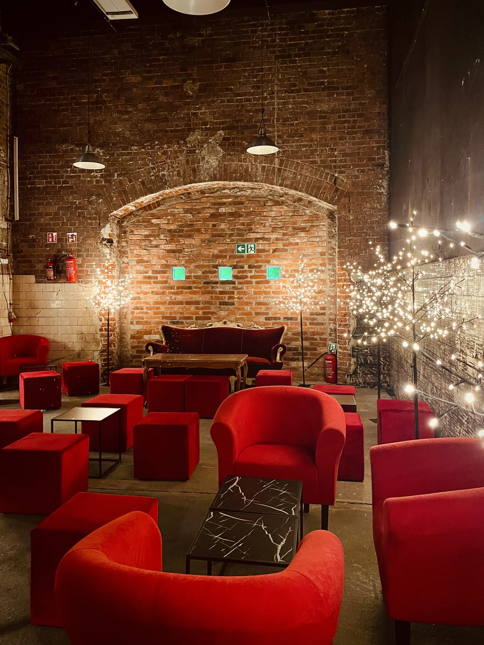 aranzacja strefa wypoczynku mikolajki swieta czarne stoliki czerwone fotele eventy wypozyczalnia dekoracji rent4events rent4wedding wroclaw - Wypożyczalnia Ślubna Czerwony chillout i Drzewka Led. Industrialna aranżacja eventowa z ceglaną ścianą w tle. Główne elementy to elegancka czerwona sofa, okrągłe fotele z aksamitną tapicerką oraz liczne sześcienne pufy w głębokim czerwonym odcieniu. Dekoracyjne drzewka LED z delikatnym światłem dodają klimatu, kontrastując z surowością loftowej przestrzeni. Czarny stolik z marmurowym wzorem dopełnia elegancję scenografii. Wypożyczalnia dekoracji na ślub, wesele i eventy we Wrocławiu - rent4events.pl wypożycz wrażenia, rent4weddings.pl zachowaj wspomnienia. Fot. Martyna.