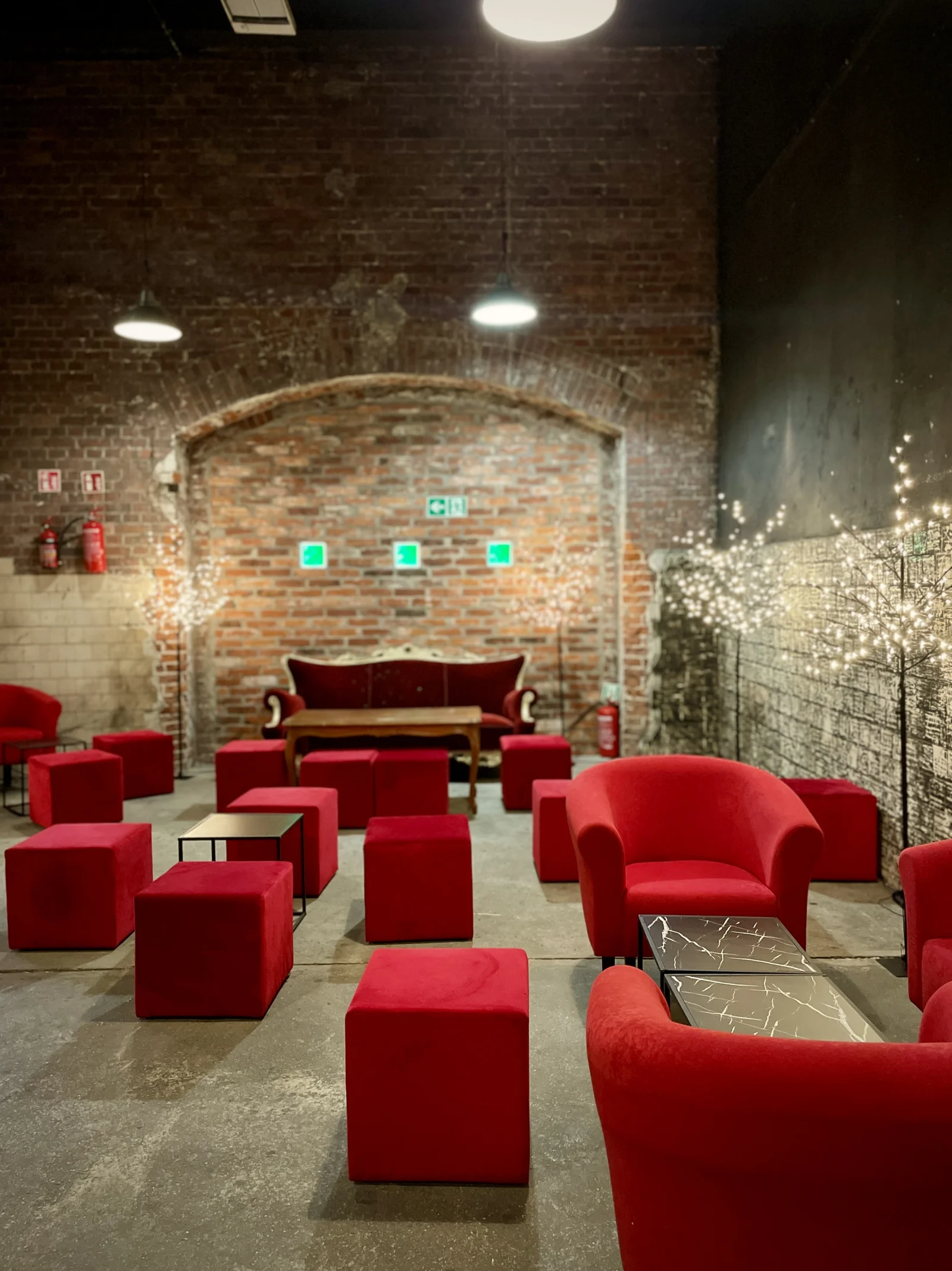 aranzacja strefa wypoczynku mikolajki swieta czarne stoliki czerwone pufy eventy wypozyczalnia dekoracji rent4events rent4wedding wroclaw - Wypożyczalnia Ślubna Czerwony chillout i Drzewka Led. Industrialna przestrzeń eventowa z ceglaną ścianą w tle. W centrum znajduje się elegancka czerwona sofa, otoczona licznymi czerwonymi sześciennymi pufami i okrągłymi fotelami o aksamitnej tapicerce. Dekoracyjne drzewka LED z delikatnym, białym oświetleniem podkreślają klimat wnętrza. Czarny, nowoczesny stolik z marmurowym wzorem dodaje elegancji przestrzeni. Surowy betonowy podkład oraz oświetlenie loftowe tworzą wyjątkową industrialną scenografię. Wypożyczalnia dekoracji na eventy, wesela i śluby we Wrocławiu – rent4events.pl wypożycz wrażenia, rent4weddings.pl zachowaj wspomnienia. Fot. Martyna.