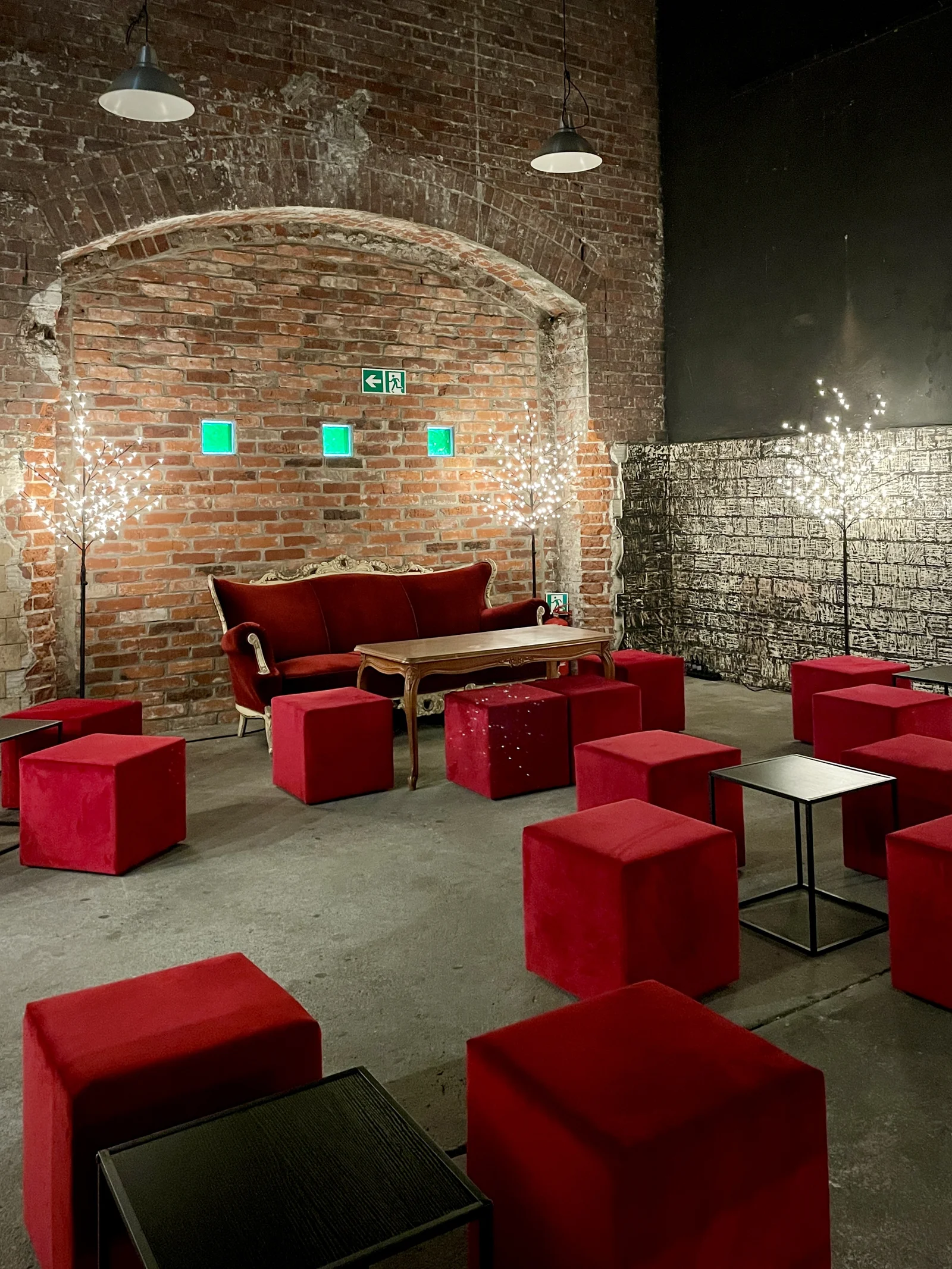 aranzacja strefa wypoczynku mikolajki swieta czerwona sofa czerwone pufy eventy wypozyczalnia dekoracji rent4events rent4wedding wroclaw - Wypożyczalnia Ślubna Czerwony chillout. Stylowa industrialna scenografia eventowa z ceglaną ścianą w tle. Centralnie umieszczona elegancka, czerwona sofa w otoczeniu licznych tapicerowanych kostek siedziskowych w kolorze czerwonym. Całość dopełniają drewniany stolik retro oraz dekoracyjne podświetlane drzewka LED, dodające przytulnego klimatu. Oświetlenie sufitowe w stylu loftowym oraz surowe, betonowe podłoże podkreślają industrialny charakter aranżacji. Wypożyczalnia dekoracji i scenografii eventowej na ślub, wesele oraz eventy we Wrocławiu – rent4events.pl i rent4weddings.pl - rent4events.pl wypożycz wrażenia, rent4weddings.pl zachowaj wspomnienia. Fot. Martyna.