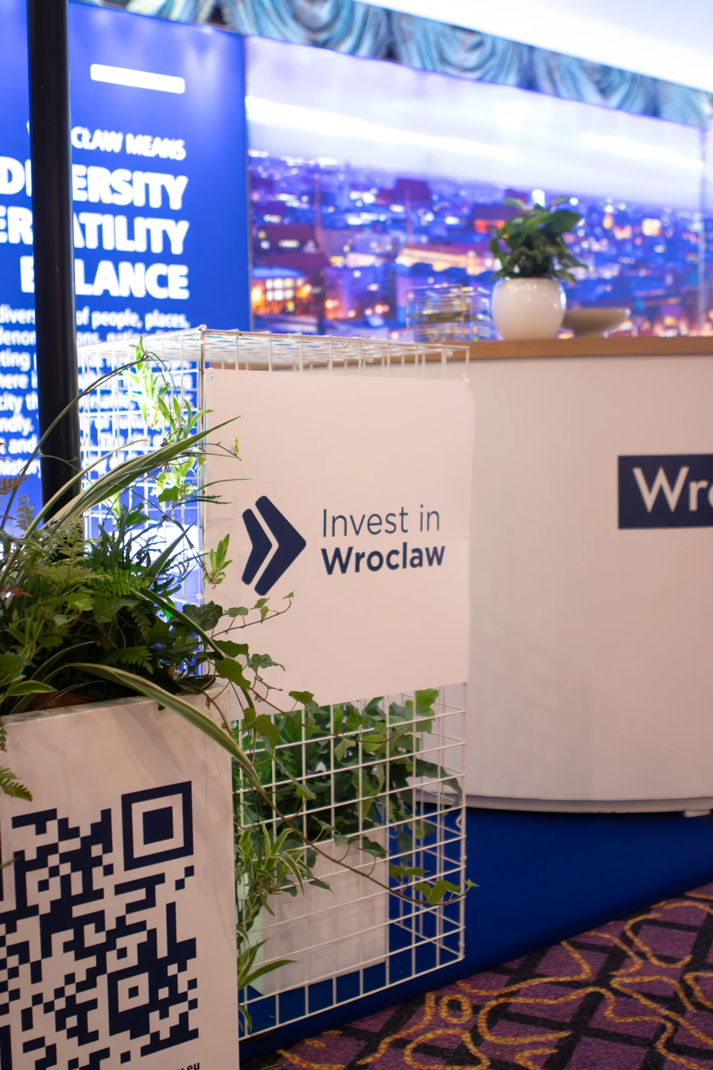 przetrzen eventowa slupek z kraty recepcja eventy wypozyczalnia dekoracji rent4events rent4wedding wroclaw - Wypożyczalnia Ślubna Stoisko targowe z brandingiem „Invest in Wrocław” przygotowane przez wypożyczalnia rent4events.pl i rent4weddings.pl to idealna scenografia na targi czy konferencje. Białe stelaże z dekoracjami roślinnymi, nowoczesne tablice, QR code oraz panorama miasta tworzą elegancką i funkcjonalną przestrzeń. Dekoracje i scenografia dostępne w wypożyczalni podnoszą prestiż każdej imprezy. Dla organizatorów eventów i uroczystości ślubnych we Wrocławiu rent4events.pl i rent4weddings.pl oferują kompleksowe rozwiązania - rent4events.pl wypożycz wrażenia, rent4weddings.pl zachowaj wspomnienia. Fot. @karolinakiec_fotografia.