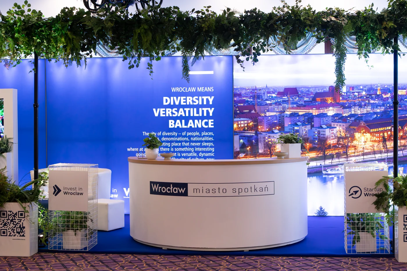 przetrzen eventowa stoisko biale meble targi eventy wypozyczalnia dekoracji rent4events rent4wedding wroclaw - Wypożyczalnia Ślubna Stoisko targowe z brandingiem „Wrocław miasto spotkań" i „Invest in Wrocław” przygotowane przez wypożyczalnia rent4events.pl i rent4weddings.pl to idealna scenografia na targi czy konferencje. Biała recepcja oraz stelaże z dekoracjami roślinnymi tworzą elegancką i funkcjonalną przestrzeń. Dekoracje i scenografia dostępne w wypożyczalni podnoszą prestiż każdej imprezy. Dla organizatorów eventów i uroczystości ślubnych we Wrocławiu rent4events.pl i rent4weddings.pl oferują kompleksowe rozwiązania - rent4events.pl wypożycz wrażenia, rent4weddings.pl zachowaj wspomnienia. Fot. @karolinakiec_fotografia.