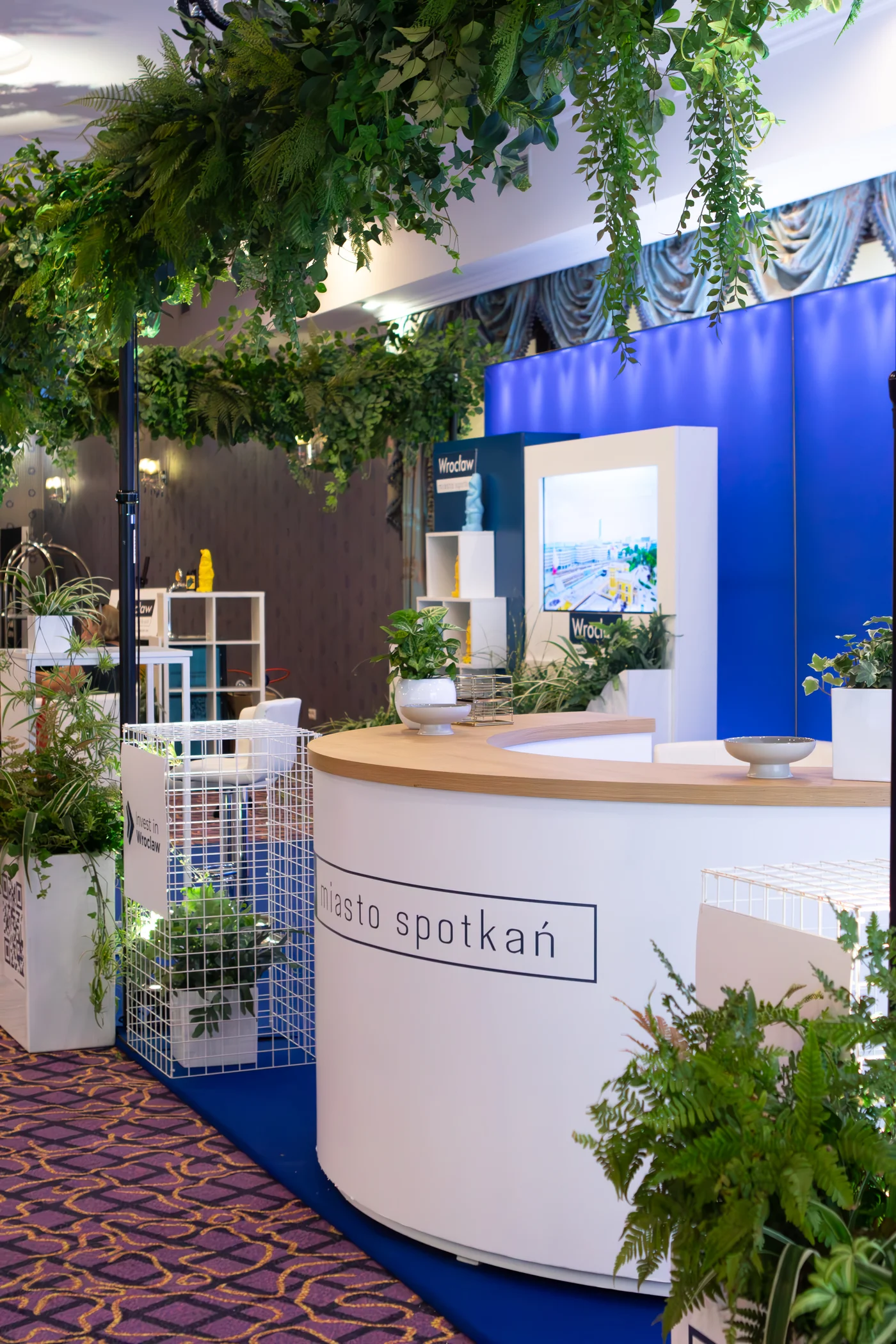 przetrzen eventowa stoisko recepcja eventy wypozyczalnia dekoracji rent4events rent4wedding wroclaw - Wypożyczalnia Ślubna Stoisko eventowe „Wrocław – miasto spotkań” z dekoracjami od wypożyczalni rent4events.pl i rent4weddings.pl to nowoczesna scenografia na targi i konferencje. Elegancka lada, białe stelaże, bujne zielone dekoracje wiszące i stojące tworzą wyjątkową atmosferę. Połączenie roślin, nowoczesnych donic i ekranów multimedialnych sprawia, że każda przestrzeń eventowa staje się przyjazna i stylowa. Wypożyczalnia rent4events.pl oferuje dekoracje dopasowane do potrzeb zarówno eventowych, jak i ślubnych, gwarantując niezapomniane wrażenia - rent4events.pl wypożycz wrażenia, rent4weddings.pl zachowaj wspomnienia. Fot. @karolinakiec_fotografia.