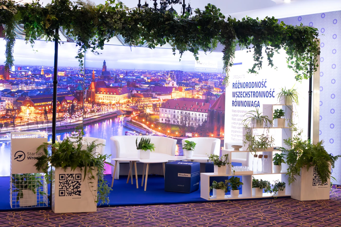 przetrzen eventowa stoisko targowe biale meble eventy wypozyczalnia dekoracji rent4events rent4wedding wroclaw - Wypożyczalnia Ślubna Stoisko eventowe „Wrocław – miasto spotkań” przygotowane przez wypożyczalnię rent4events.pl / rent4weddings.pl. Eleganckie białe meble, zielone rośliny w donicach, wiszące dekoracje i tło z panoramą miasta tworzą wyjątkową scenografię. Wypożyczalnia dekoracji oferuje kompleksowe wyposażenie i aranżacje, które podnoszą prestiż każdej uroczystości. Dla organizatorów ślubów, wesel i eventów we Wrocławiu i okolicach rent4events.pl i rent4weddings.pl to gwarancja stylu i jakości - rent4events.pl wypożycz wrażenia, rent4weddings.pl zachowaj wspomnienia. Fot. @karolinakiec_fotografia.